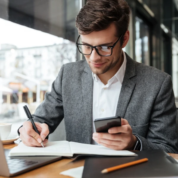 Homem usando um celular e fazendo anotações em um caderno